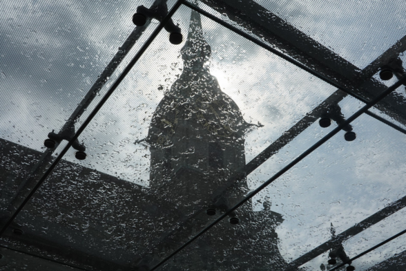 Glasdach, getragen von Metallstreben, darauf Wassertropfen, darüber Kirchturm und Wolen - Sonne hinter'm Kirchturm