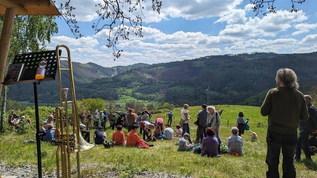 Eine Rast der Wandergruppe 2024 mit Blick ins Loquitztal. Pfarrer Gindler steht vorn rechts, auf der Wiese die WanderInnen. Links steht eine Posaune und ein Notenständer.