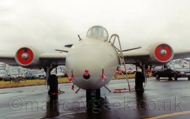 Head-on view of a twin-engined military jet bomber plane parked on black tarmac facing the camera.
The plane is mostly sandy-brown, with a grey belly.
The engines are buried in the wings, with red plates over the intakes, and the main landing gear is attached to the side of the engine pods.
The bubble canopy over the cockpit is offset to the left of the plane, with a removeable ladder attached for the crew to access the plane.
Red and white ribbons are attached to the covers of various sensors on the planes nose.
Puddles on the black tarmac suggest that it has recently been raining.
A field full of parked cars is in the background, stretching off into the distance, under flat grey sky.