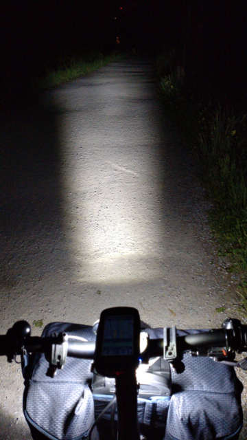 Nächtlicher Blick auf den Lenker meines Brompton Electric G-Line mit Garmin GPS und darunter einer Ravemen "zum Gesehen werden" Leuchte. Man sieht gut die große graue Fronttasche und den Lichtkegel des unter der Fronttasche montierten Scheinwerfers.