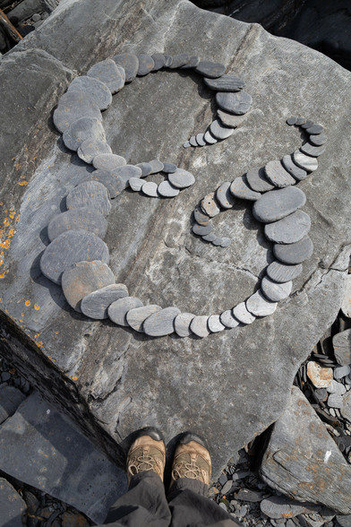 An ampersand formed of overlapping rounded slate stones. On a bigger, flat, rock. The photographer's feet are at the bottom of the frame.
