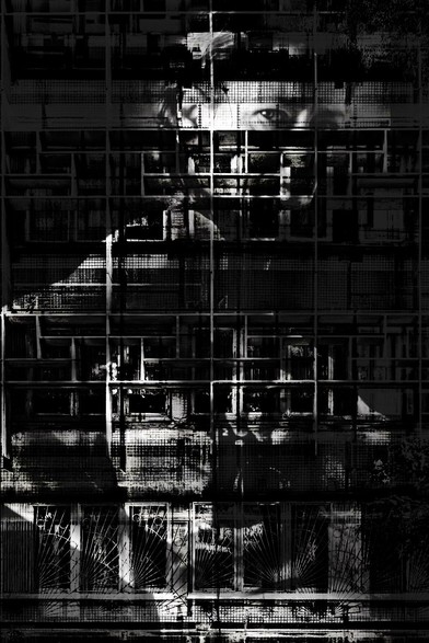 Black and white photograph, portrait format, multiple exposure. Self-portrait with architectural elements (Brutalist façade with many windows). Through a dark grid of windows dominated by the horizontal and vertical lines of the façade, the shadowy body and head of a woman are discernible. She has turned her back to the camera, but her head is turned so that it looks at the viewer as if through a window. She is reflecting herself into a mirror. The overall impression is gloomy, the subject's gaze critical and inquisitive; the architectural elements act like a prison in front of the body.