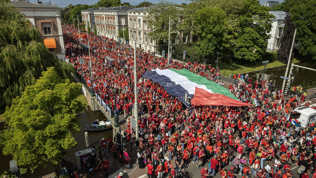 Een enorme stoet demonstranten, veelal in rood gekleed, vormen een lange rode 'streep' door de straten van Den Haag, terwijl ze een grote Palestijnse vlag horizontaal boven hun hoofden meedragen.