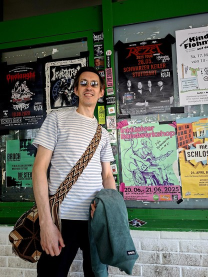 Ein großer Mann mit dunklen Haaren und Sonnenbrille steht vor einer plakatierten Wand. Rechts neben ihm das Plakat des Inklusiven Bandworkshops