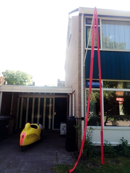 Velomobile parked outside a house in the Netherlands with a long red line of knitted scarf dangling from the flagpole.
