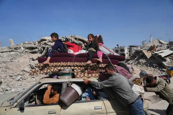 People push a car full of people, stacked with mattresses with children sitting on top.