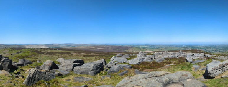 Blue sky above a distant, hazy horizon.  Rocky outcrops nearby.  Between, a moorland hilltop saddle gives way to moorland hills to the left, descending to mixed rural and increasingly settled land to the right.