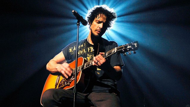 Chris Cornell, el icónico vocalista de Soundgarden y Audioslave, está sentado tocando una guitarra acústica. Está iluminado desde arriba por un foco brillante que crea rayos de luz a su alrededor. Viste una camiseta oscura con un diseño gráfico y se concentra en su interpretación. El fondo es oscuro y la escena está ambientada en un escenario.