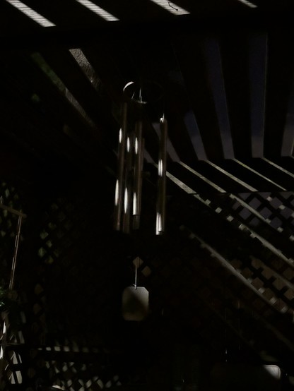 Black-and-white night time photo of wind chimes illuminated by a street light filtered through slats of wood