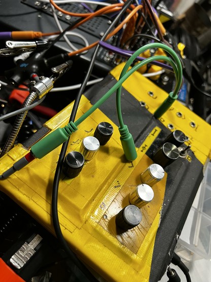 The image shows a close-up view of an electronic device with a yellow and black casing. The device features several silver knobs, likely for adjusting settings, and is connected to various cables. Two green cables with connectors are prominently visible, one plugged into a jack on the device. The background is filled with additional cables and electronic components, suggesting a complex setup. The device appears to be part of a larger system, possibly for audio or signal processing, given the presence of audio jacks and the intricate wiring. The image captures the detailed arrangement of the device and its connections, highlighting the complexity of the setup.