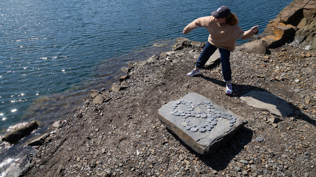 An ampersand on a big square rock. A woman stands next to the rock, skimming stones on the still water of the flooded quarry.