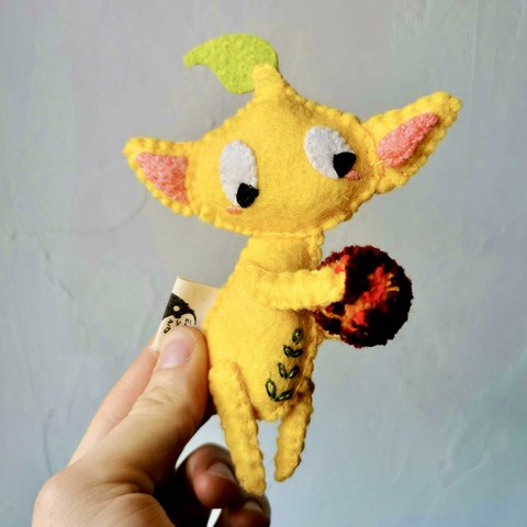 A handmade felt pikmin plushie held in front of a white wall. 
The pikmin is yellow, with big ears and a little leaf on top of its head. Little leaves are embroidered on it’s belly and it is holding a pompon that looks like a bombrock.