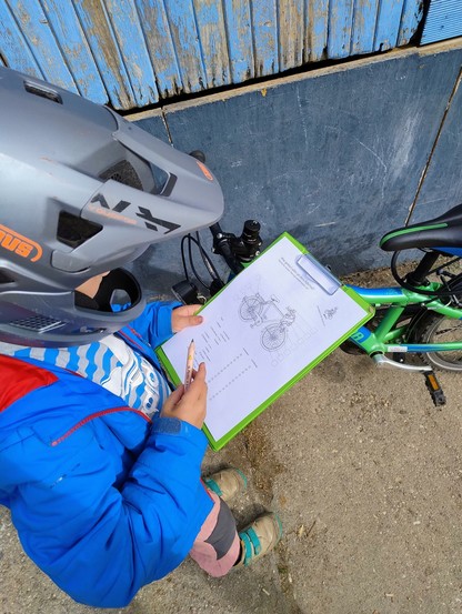 Das Bild zeigt ein Kind mit einem Sturzhelm in blauer Jacke, das ein Schreibbrett mit einem Papier, auf dem ein Fahrrad und Ankreuzfelder zu sehen sind und einen Stift in der Hand hält und vor einem Fahrrad steht.