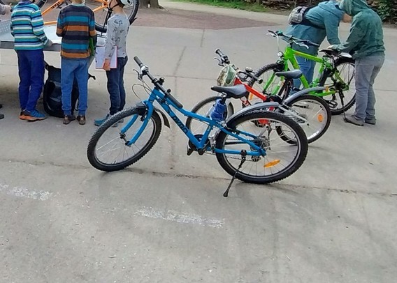 Das Bild zeigt zwei Kinder in blau/grau/oranger Kleidung und eine erwachsene Person in blau, die neben oder an drei Fahrrädern in blau, rot und grün stehen.