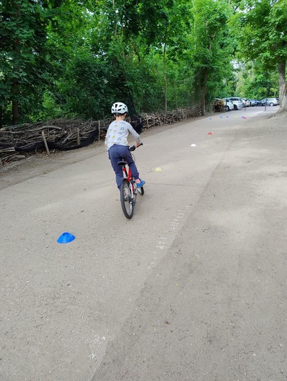 Das Bild zeigt ein Kind mit weißem Helm, hellblau gemustertem Pullover und blauer Hose von hinten, welches auf einem roten Fahrrad auf einem Weg um bunte kleine Hütchen Slalom fährt.