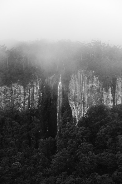 black and white waterfall on a foggy rainy day. Trees at the bottom, cliffs with the waterfall in the middle, trees at the top partially covered with the fog and then foggy sky.