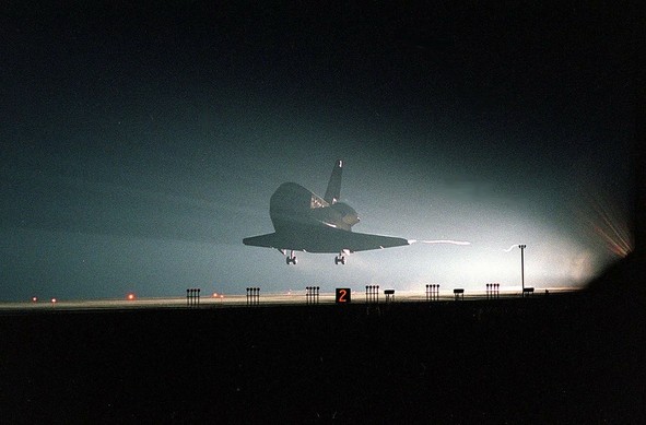 A space shuttle is landing at night, silhouetted against a backdrop of light. Ground lights and a numbered marker are visible in the foreground.