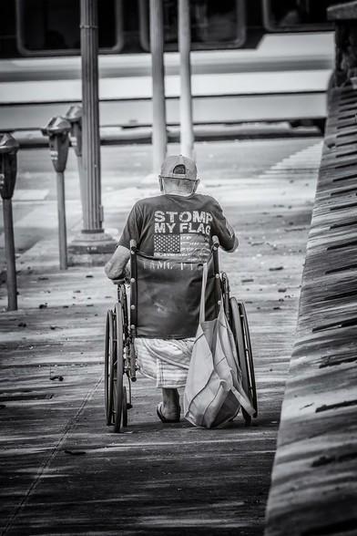 Man (most likely a veteran) in a wheelchair going down a sidewalk in Honolulu's Chinatown. On the back of his t-shirt is an American flag with the caption "Stomp My Flag" above it.