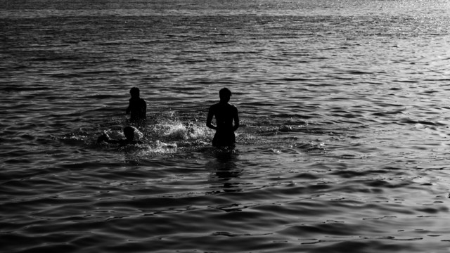 The image shows three people in silhouette, standing and swimming in a body of water. The scene is backlit, casting the figures in shadow and emphasizing the ripples on the water's surface. The photograph has a tranquil and serene quality, capturing a moment of leisure.