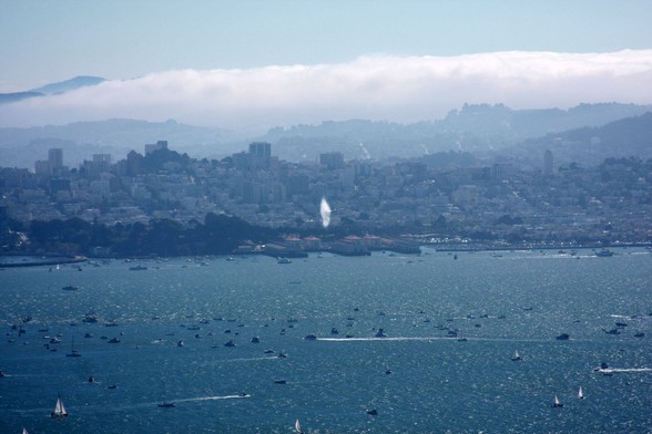 With the San Francisco Bayfront behind it, a Blue Angels jet creates a vapor cone during supersonic flight at low altitude over the water and the many boats below.