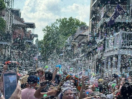 Photo of street in the French Quarter of New Orleans. On either side are 2 and 3 story buildings, many with balconies and galleries, typical of the historic district.   Between and in foreground, a crowd of people fill the street, many colorfully dressed, some hold up smart-phones taking photos or videos, others holding up various bubble machines and bubble blowers.  The whole scene is seen through thousands of floating soap bubbles filling the air. Photo by Jenny Campbell.