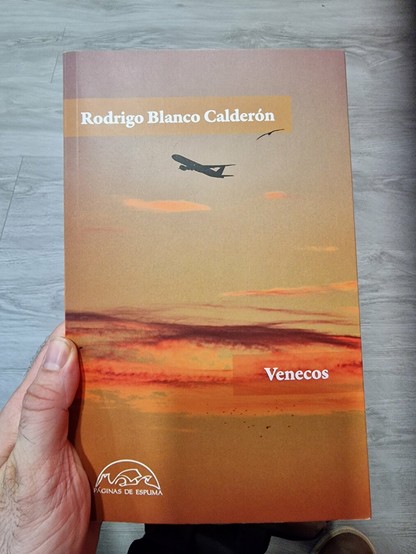 Una mano sostiene un libro de tapa blanda. La portada es de color marrón rojizo, con una imagen de un avión volando sobre un cielo anaranjado al atardecer. El título del libro, "Venecos", está escrito en la parte inferior. En la parte superior, aparece el nombre del autor: "Rodrigo Blanco Calderón". En la parte inferior de la portada, hay un logotipo que dice "Páginas de Espuma".