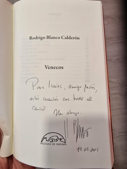La imagen muestra la página de dedicatoria de un libro titulado "Venecos", escrito por Rodrigo Blanco Calderón. La página contiene una dedicatoria manuscrita a un "Isaías, amigo lector", deseándole que disfrute de los cuentos. La dedicatoria está firmada y fechada el 19 de mayo de 2025, con el logo de "Páginas de Espuma", la editorial.