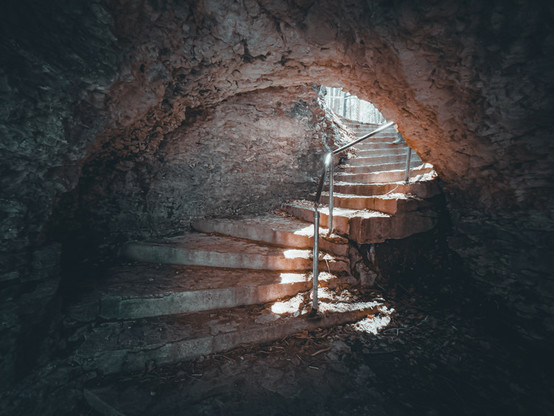 The image depicts a staircase that leads to an underground area, possibly a cave or a tunnel. The stairs are made of stone and appear to be old and rusted. There is a metal railing on the side of the staircase for support. Above the staircase, there is a natural light source coming through, illuminating the space. The overall atmosphere of the image conveys an eerie or mysterious feeling due to the underground location and the aged appearance of the stairs.