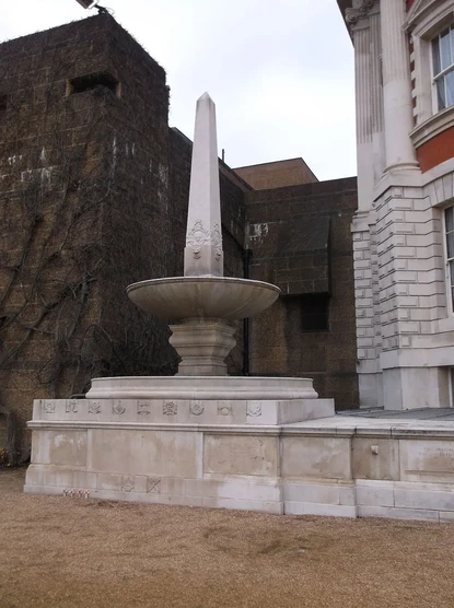 The image features an obelisk-style fountain, positioned near what appears to be a historical building. The fountain has a circular basin and a tall, tapering structure on top resembling an obelisk. The background includes a wall covered in vines or ivy, alongside a more modern structure. The scene is likely set in a courtyard or garden area of the building.

Image Credits: Wikimedia / Harry Mitchell / CC BY 4.0