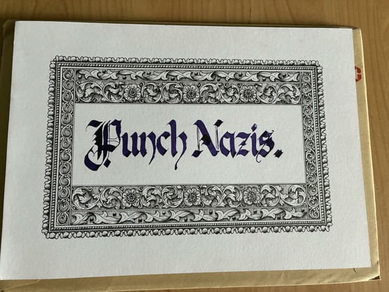 Ein Foto eines weißen Blatt A4. Darauf am Rand eine gemalte Bordüre? in einem klassischen Stil.
Innen kaligrafiert die Worte „Punch Nazis.“