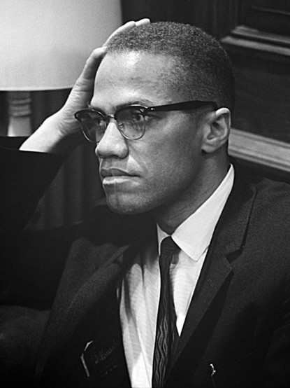 Malcolm X im März 1964, im dunklen Anzug, weißem Hemd mit Krawatte, den Kopf mit dem Rechten Arm abstützend