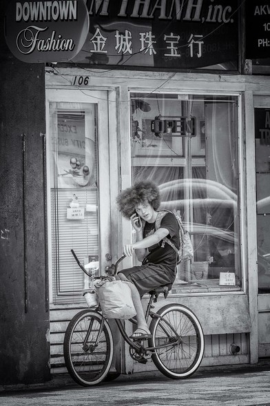 Young man with big hair sits on a bicycle underneath a sign for a clothing store ("Downtown Fashion") in Honolulu's Chinatown.
