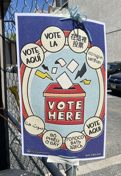 A cartoon-style sign shows a red ballot box marked in white letters "Vote Here" -- a little card is going into it, surrounded with comic book lightning bolts (the power of the vote?).  Around this are red and white bands, like a bullseye, with four small bubbles above and below the ballot box giving the same info in a variety of languages (Spanish, French, Italian? Mandarin, Arabic, Thai, and several less familiar writing systems).  The sign is hanging on a chain-link fence outside the parking lot of a polling place.