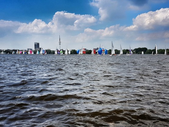 A wide shot of the lake in Hamburg centre filled with many sailboats with colourful sails. In the background is a city skyline under a blue sky with white clouds.