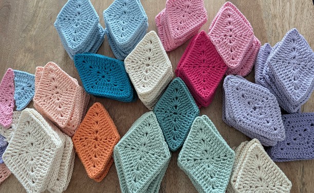 Picture of stacks of crocheted lozenges in various colors: cream, lilac, purple, peach, orange, pink, fuchsia, mint, dark green, blue,  petrol. All placed on a wooden table.