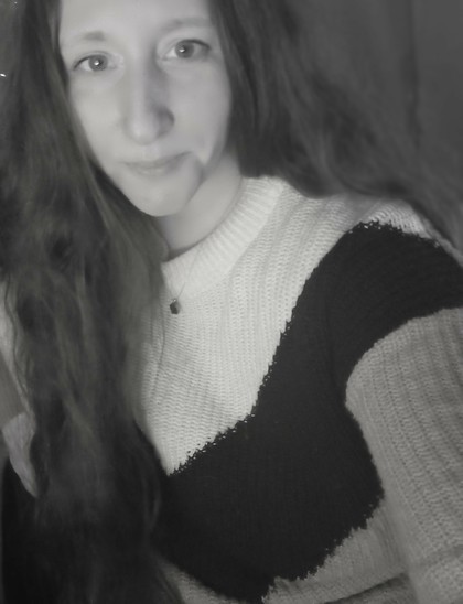 A black and white photo of a woman with long wavy hair cascading over her shoulder. She is wearing a cozy knit sweater with a bold color-block pattern consisting of light and dark contrasting sections, including a prominent diagonal dark band across the chest. Her expression is calm and slightly smiling, with a gentle gaze directed at the camera. A delicate pendant necklace rests at the neckline of her sweater. The background is softly blurred.