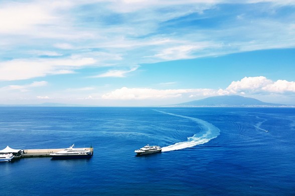 An aerial view of a deep blue ocean with two boats. One boat is docked at a pier on the left, and another boat is moving towards the right, leaving a curved white wake behind it. In the background, a large mountain rises under a partly cloudy blue sky.