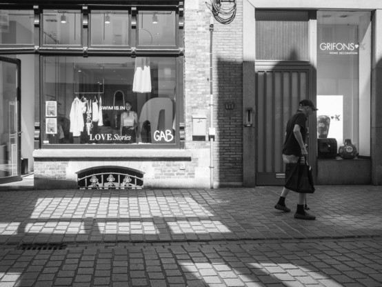 The image is a black and white photograph of a street scene. On the left, there is a storefront with large glass windows displaying clothing items, including a white dress and shirts. The window has text that reads "SWIM IS IN," "LOVE Stories," and "GAB." The building has a brick facade with a decorative metal grate at the base of the window. On the right, there is another storefront with a glass door and window, displaying home decor items, with the text "GRIFONS HOME DECORATION" visible. A man is walking on the sidewalk, wearing a dark cap, a black t-shirt, shorts, and black shoes, carrying a black bag. The ground is paved with cobblestones, and shadows from the buildings and the man are cast on the ground, creating a pattern.
