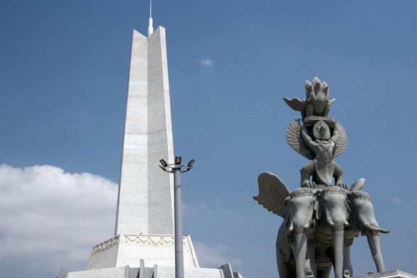 Photo shows a large stele with an elaborate statue in front. It is made of light-coloured stone and concrete. The background is a light blue sky with a few scattered clouds. The stele is clearly divided in three main parts, with a single spire in three parts on top. Probably with a lightning rod. The statue has a three headed elephant with wing as it base. It is ridden by a Garuda, a guardian figure with a bird of prey head, wings and feet, and human torso and arms. The Garuda supports a platform with a naga statue, a three headed snake with wings. Between the stele and the statue there is a light post one thirds the height of the stele with seven powerful floodlights.