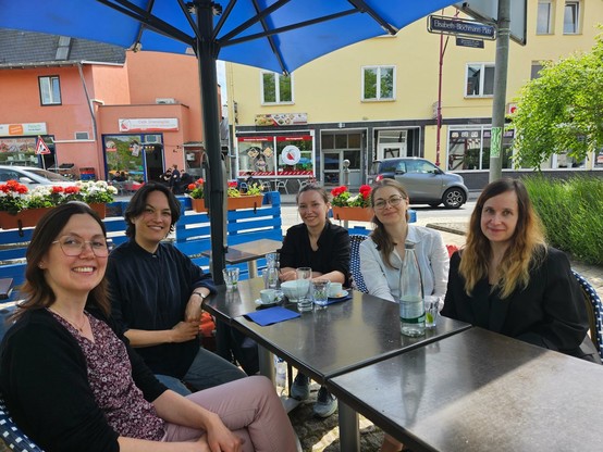 Bianca Sedmak, Sarah-Mai Dang, Pauline Junginger, Kristina Rose und Natalie Filipiak sitzen an einem Restauranttisch neben einer belebten Straße und blicken in die Kamera.