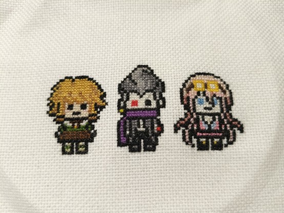 three cross stitched pixel art characters on a white weave. from left to right they are: Chihiro, who has brown hair and green and brown clothes. Gundham, who has gray hair, dark gray clothes and a purple scarf. Miu, who has pink hair and pink clothes and a pair of goggles on top of her head.