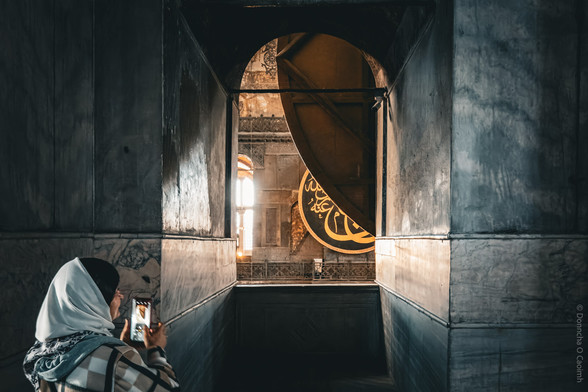 A woman in a headscarf takes a photo with her phone inside the Hagia Sophia, Istanbul, standing in a dimly lit marble corridor with a dramatic view of a large circular Islamic calligraphy medallion and arched window illuminated by sunlight; the medallion features intricate Arabic script.