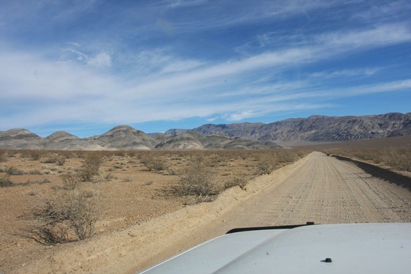 Looking over the white hood of a Land Cruiser you can see a washboarded dirt road cutting through pristine desert. Hills with interesting geology are in the background and the sky is partly cloudy.
