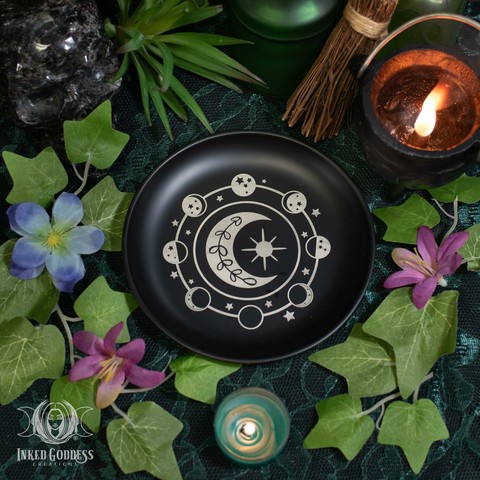 A "Moon Phase Metal Altar Dish" product is placed on a dark lace-covered surface. It's a shallow black-glazed metal bowl with moon phases and stars in a circle around a larger crescent moon and sun. Presented by Inked Goddess Creations.