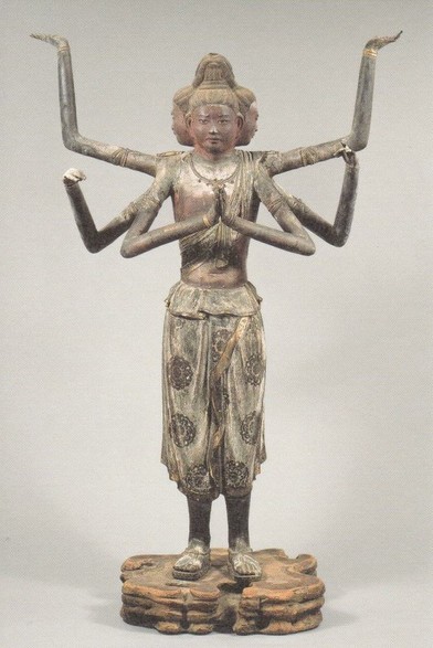 a tall  realistic sculpture of a slender figure with a 4-sided head and six arms, the arms held out in different angles, wearing baggy short pants with patterns on them, facing the viewer, against a grey background