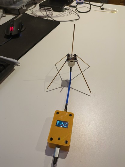 A small yellow 3D printed Meshtastic node connected to a self build ground plane antenna on a white desk. 

The antenna has 4 legs about 9cm each which are shaped like the sides of a pyramid. On the top there is a 8 cm wire pointing upwards.

In the background there a a small part of a black keyboard visible, a black mouse pad and a mouse, some cables and other stuff.