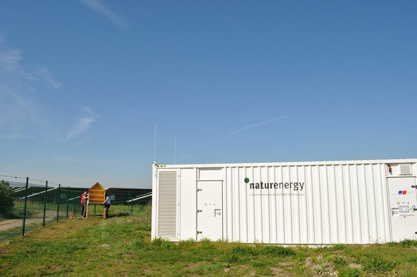 Ein weißer Container mit der Aufschrift "naturenergy – Ein Unternehmen der NATURSTROM-Gruppe" steht auf einer Wiese. Im Hintergrund montieren zwei Personen ein großes Insektenhotel an einem Zaun in der Nähe eines Solarparks.