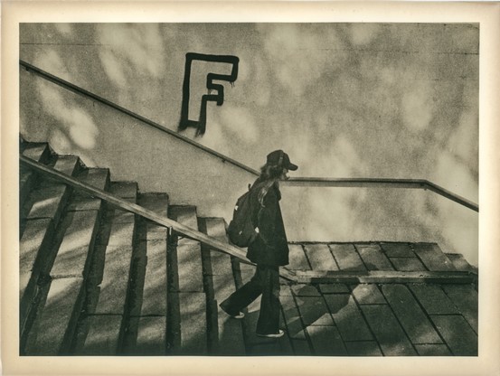 This image is a lith print photograph, characterized by its distinctive gritty texture, high contrast, and warm, often sepia-toned hues.

The scene captures a solitary figure—likely a young woman—ascending a wide, outdoor staircase. She is dressed in dark clothing, wearing a coat, a baseball cap, and a backpack. The staircase and the adjacent wall create strong diagonal lines that guide the viewer's eye through the composition. On the wall, the letter "F" is boldly spray-painted in a stylized, graffiti-like font.

Soft, dappled light filters across the scene, likely from trees casting shadows, adding a dreamlike or abstract quality. The use of lith printing enhances the mood: the shadows are deep and textured, while the highlights retain a delicate softness. The borders of the image are slightly darker and warm-toned, typical of prints on aged or fiber-based paper.