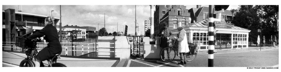 Black and white panorama photograph of two people on the sidewalk of a bridge in Zaandam, deep in conversation. One is holding their bike while the other is looking in their grocery bag. On the left a lady on a electric fatbike has just whizzed past on the bike path. In the background is the Wilhelmina Lock surrounded by various older buildings.