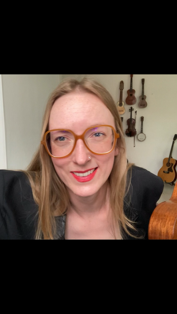Sage leans forward, smiling, holding a ukulele in one hand with a wall of instruments behind her.